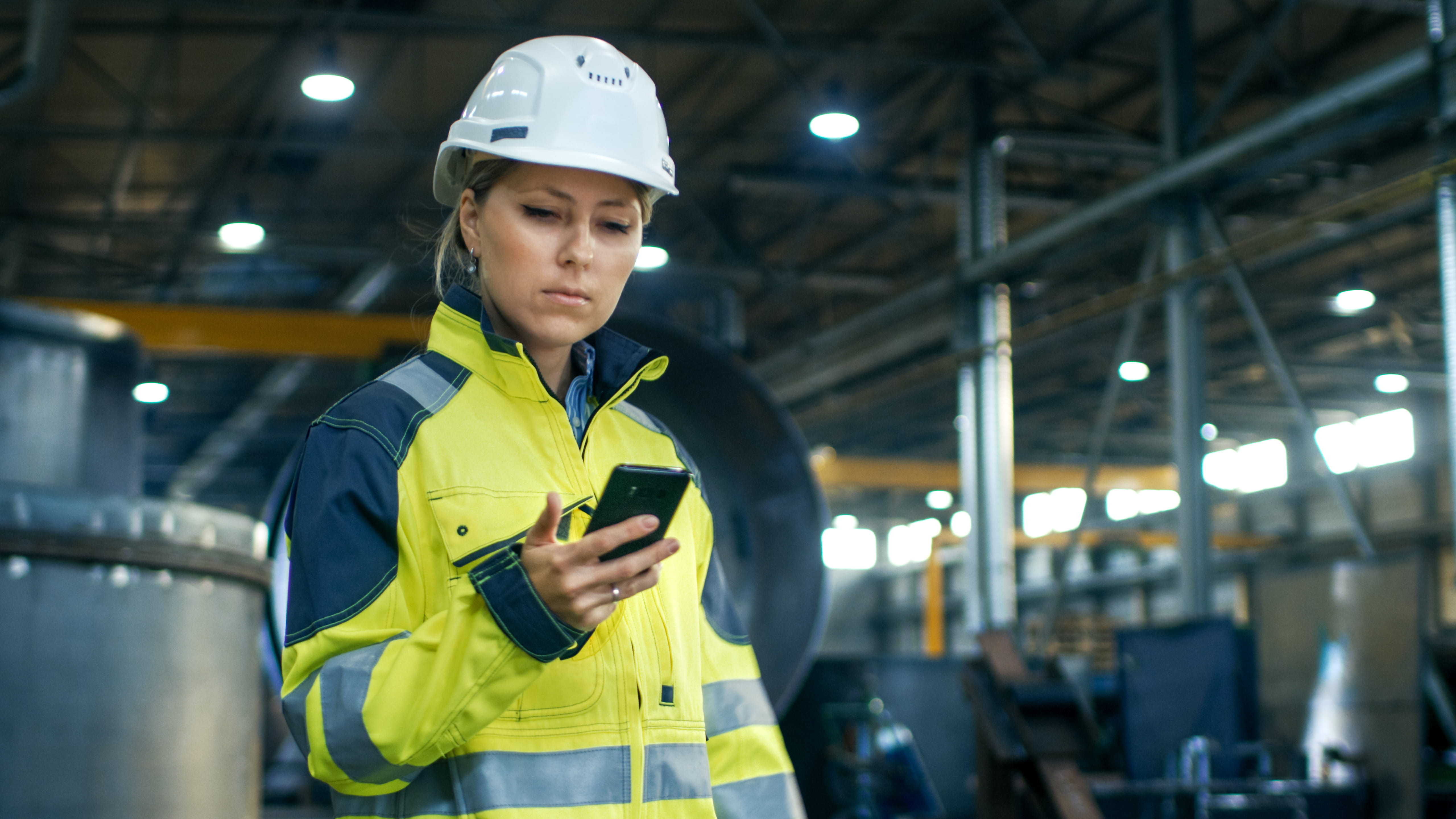 Sur le chantier, une contremaîtresse consulte l’avancement de son équipe sur son téléphone intelligent; elle porte une veste et un casque de sécurité.